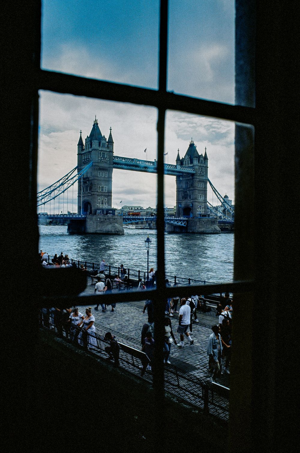 View of London's Tower Bridge and Thames River through a window frame, with tourists visible on the riverbank below.