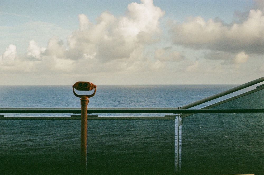 Coin-operated binoculars on a green railing overlooking a vast ocean horizon with puffy clouds in the sky.