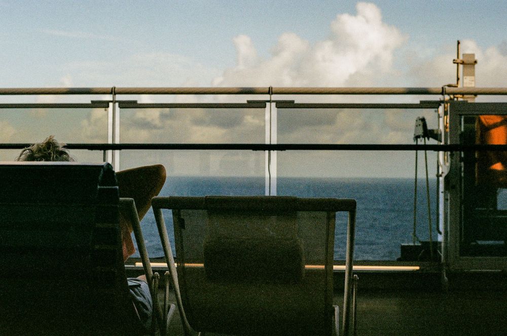 Empty chairs on a ship deck railing overlooking a calm ocean view with white clouds in the distance.