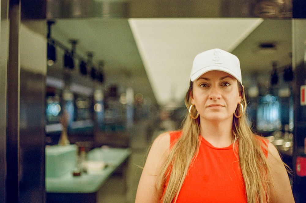 Person wearing a white baseball cap and red tank top stands in front of blurred indoor background.