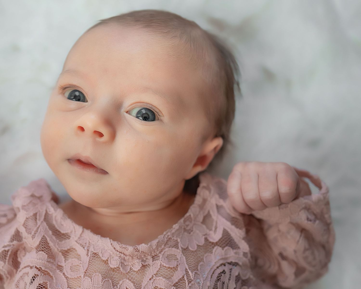 Baby wearing a delicate pink lace dress looks up with bright eyes against a soft gray background.