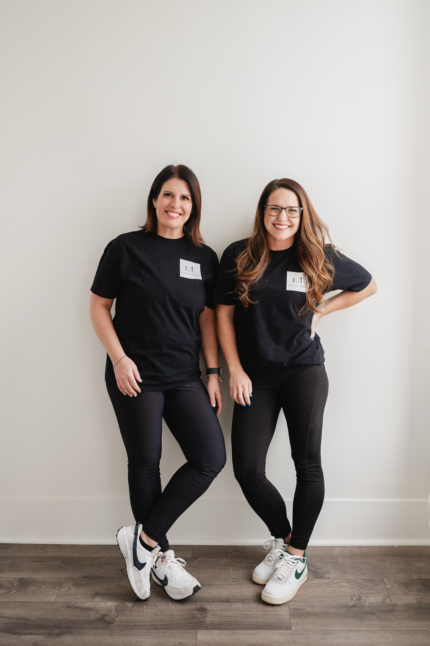 Two people in matching black outfits striking fun poses against white wall.
