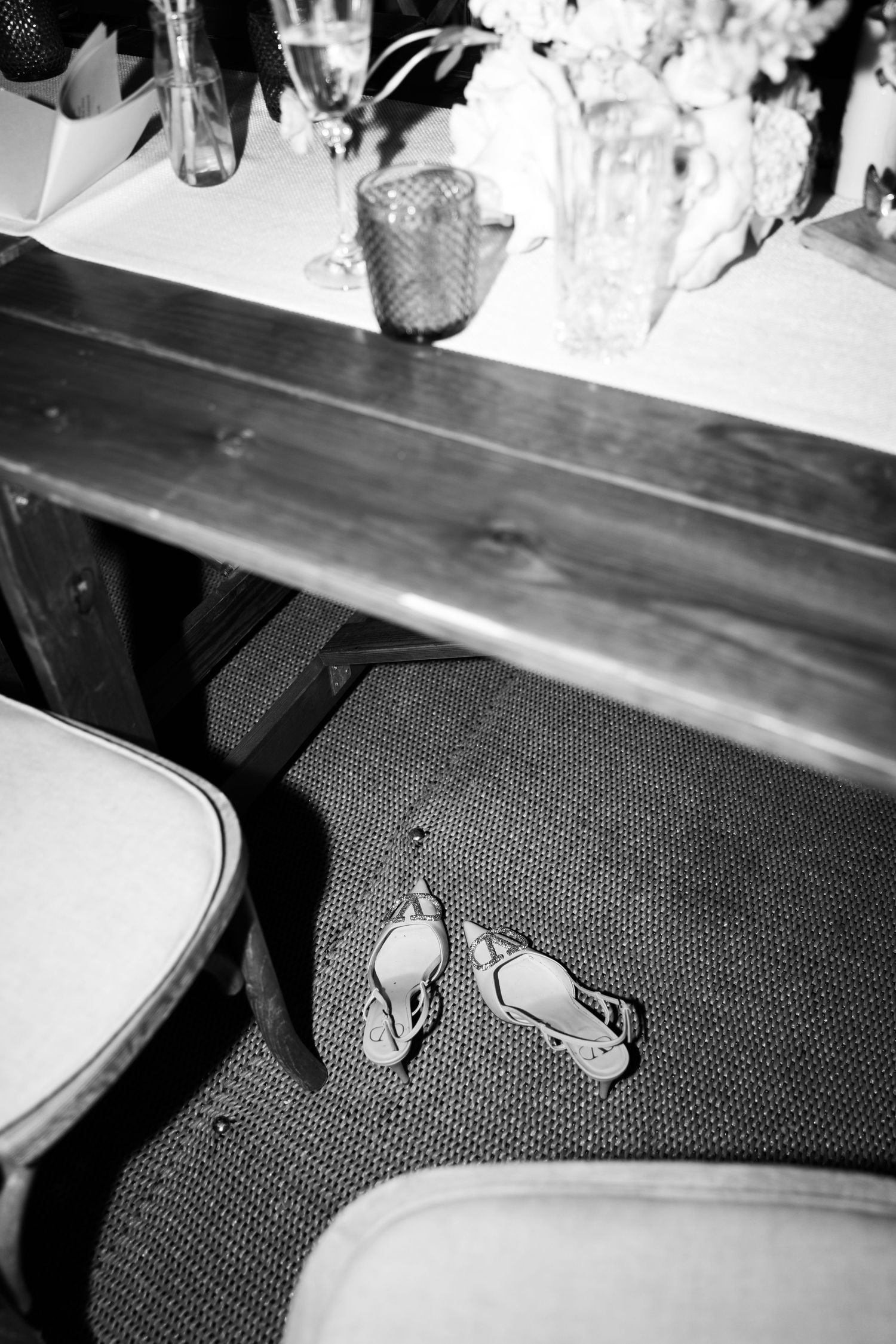 A pair of silver sandals resting on a carpeted floor beneath a wooden table in black and white.
