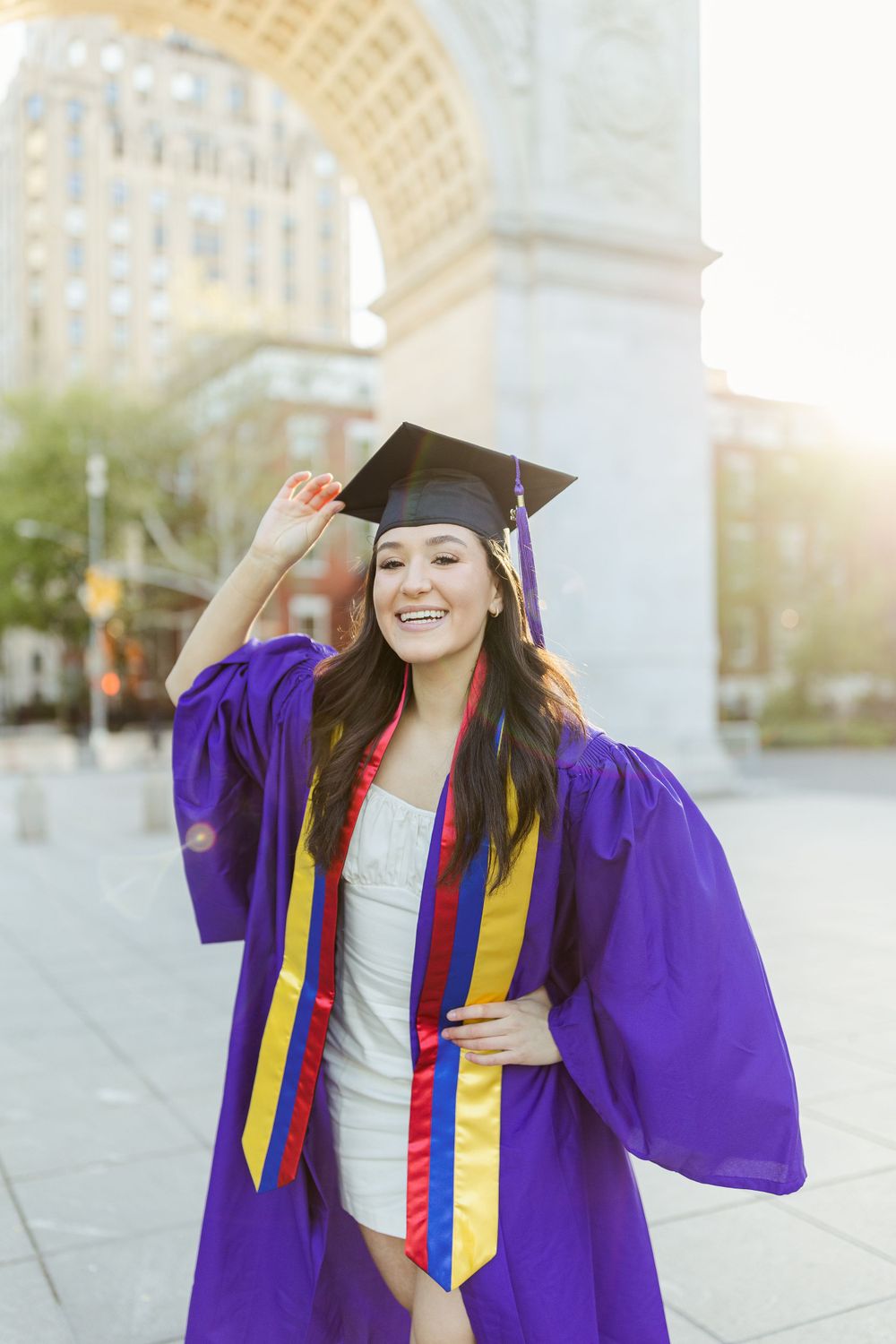 Graduate in purple and gold regalia celebrates joyfully at sunset under city arch.