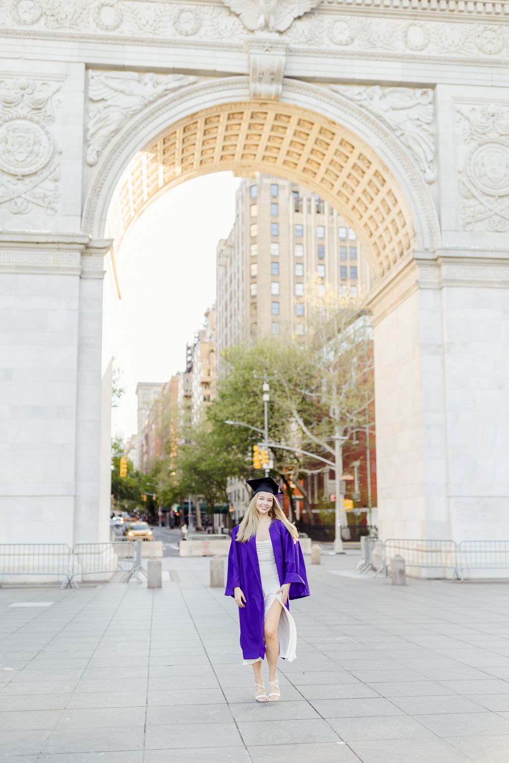 Graduate in purple commencement gown walking beneath Washington Square Arch in New York City.