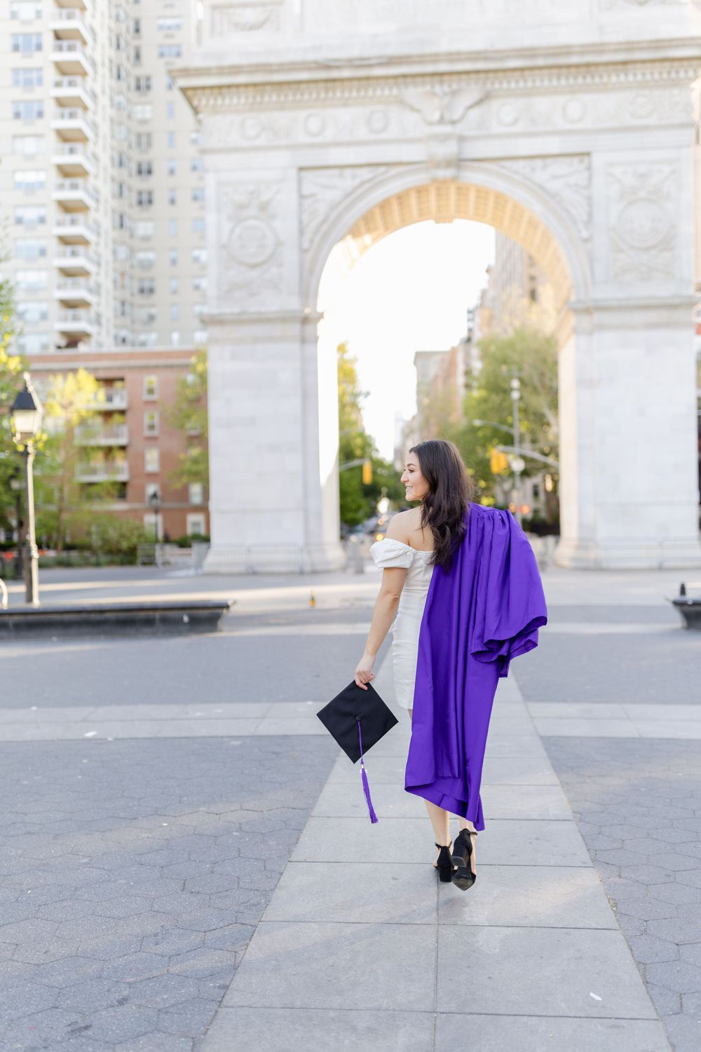 Graduate holding diploma walks near Washington Square Arch in purple graduation attire.