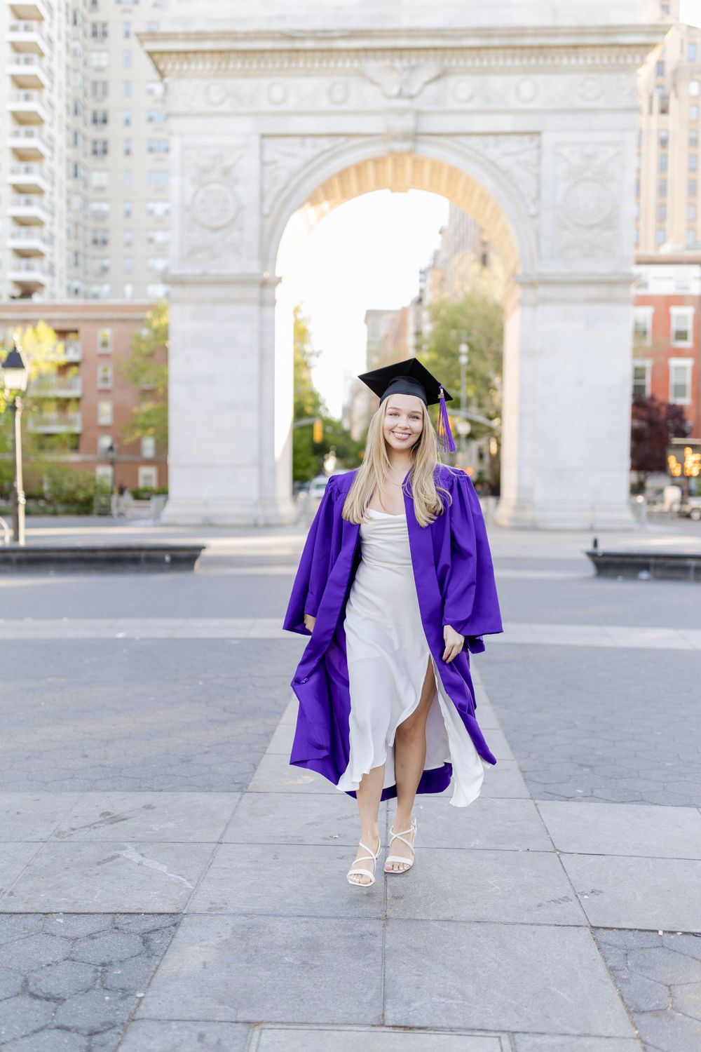 Graduate in flowing purple commencement gown walking through Washington Square Park.