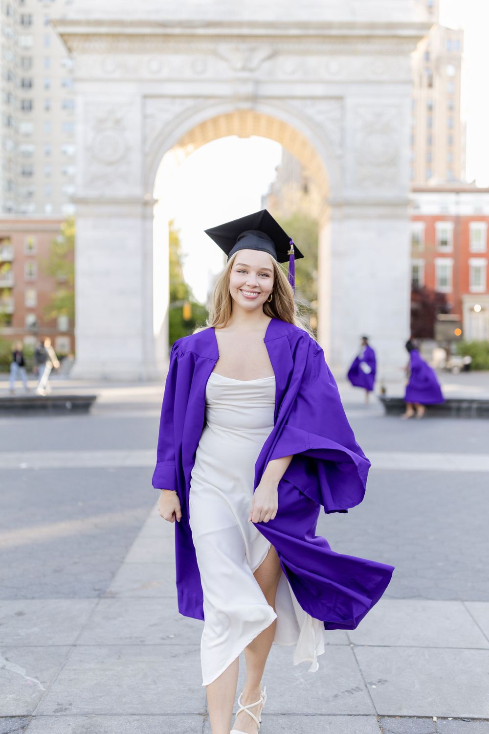 Graduate in white dress and purple graduation robe poses at Washington Square Park.