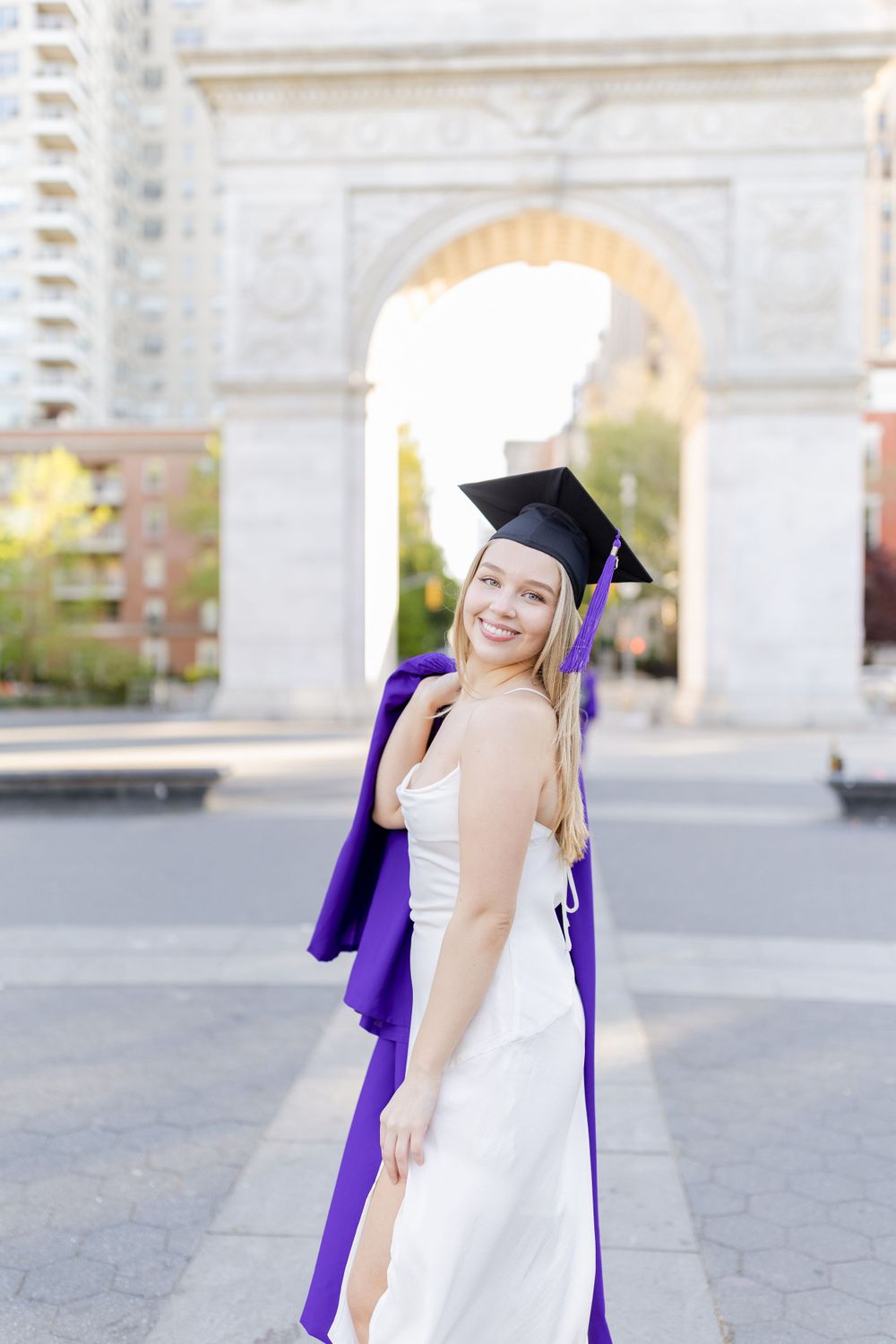 Graduate in purple commencement attire poses by historic arch in Greenwich Village.