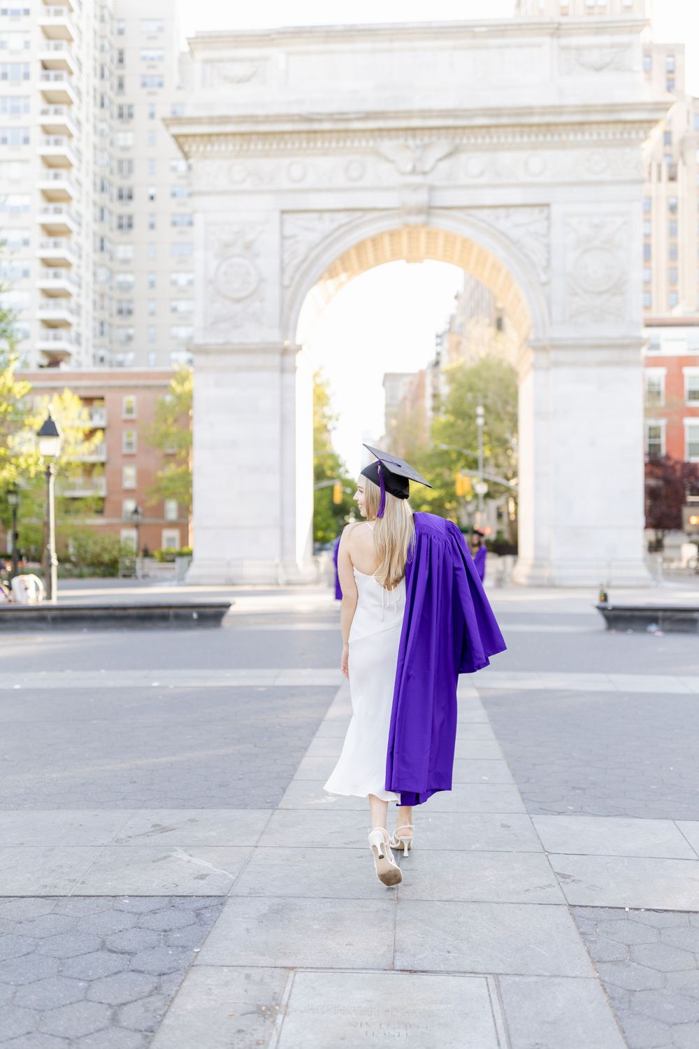 Graduate in white dress and purple graduation gown walking away from Washington Square Arch.