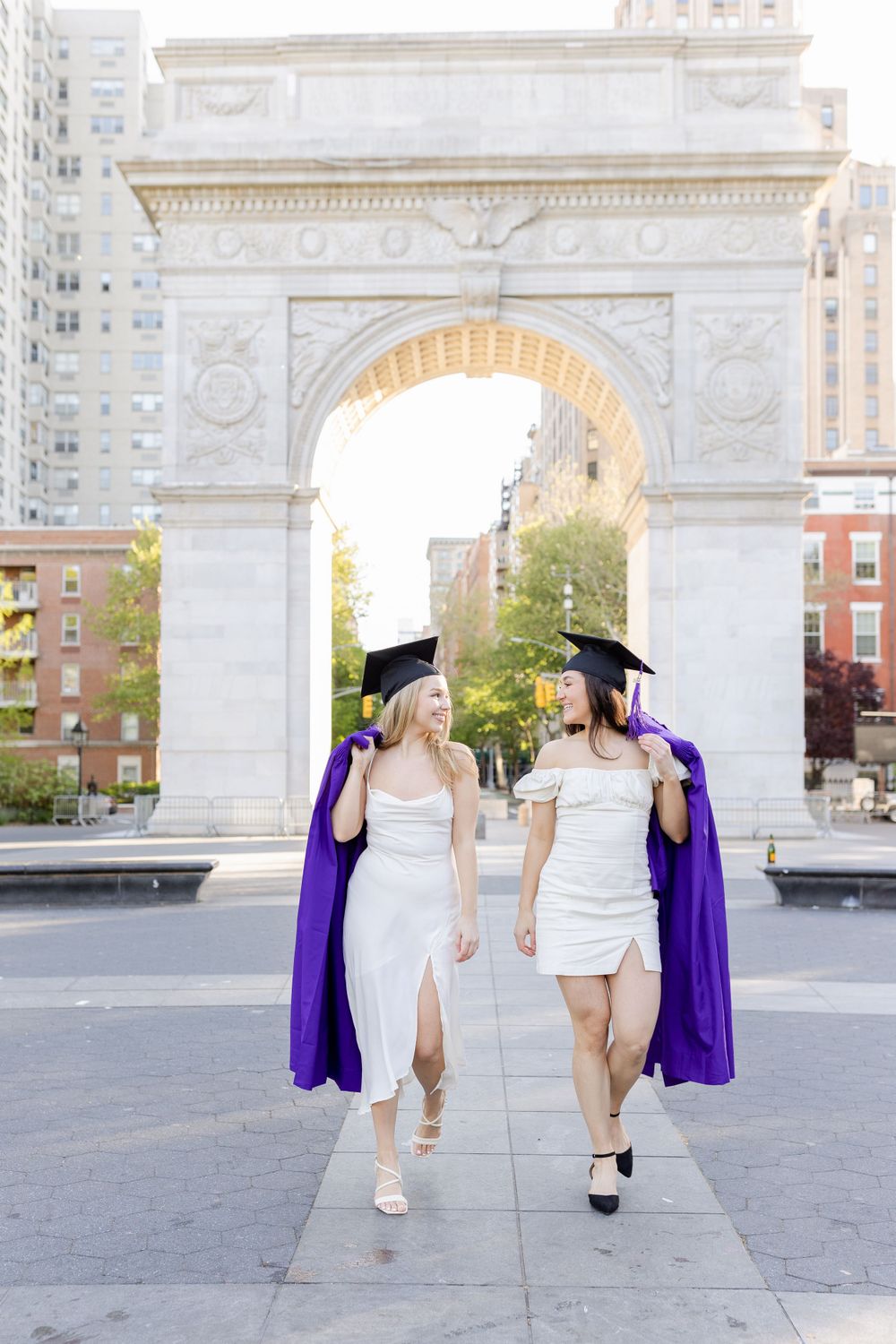 Two graduates in white dresses and purple gowns walk together under Washington Square Arch in NYC.