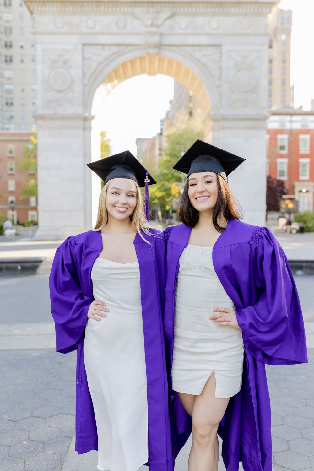 Two graduates in purple caps and gowns smile together under Washington Square Arch.