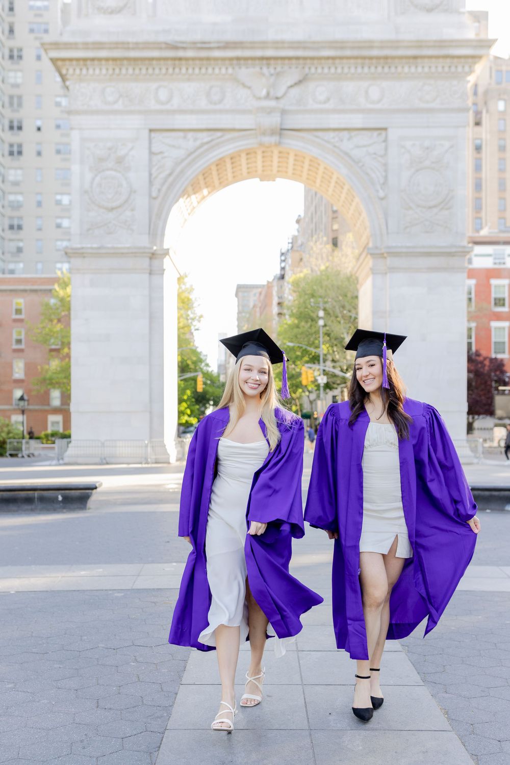 Two graduates walk and smile while wearing purple graduation robes near historic arch.
