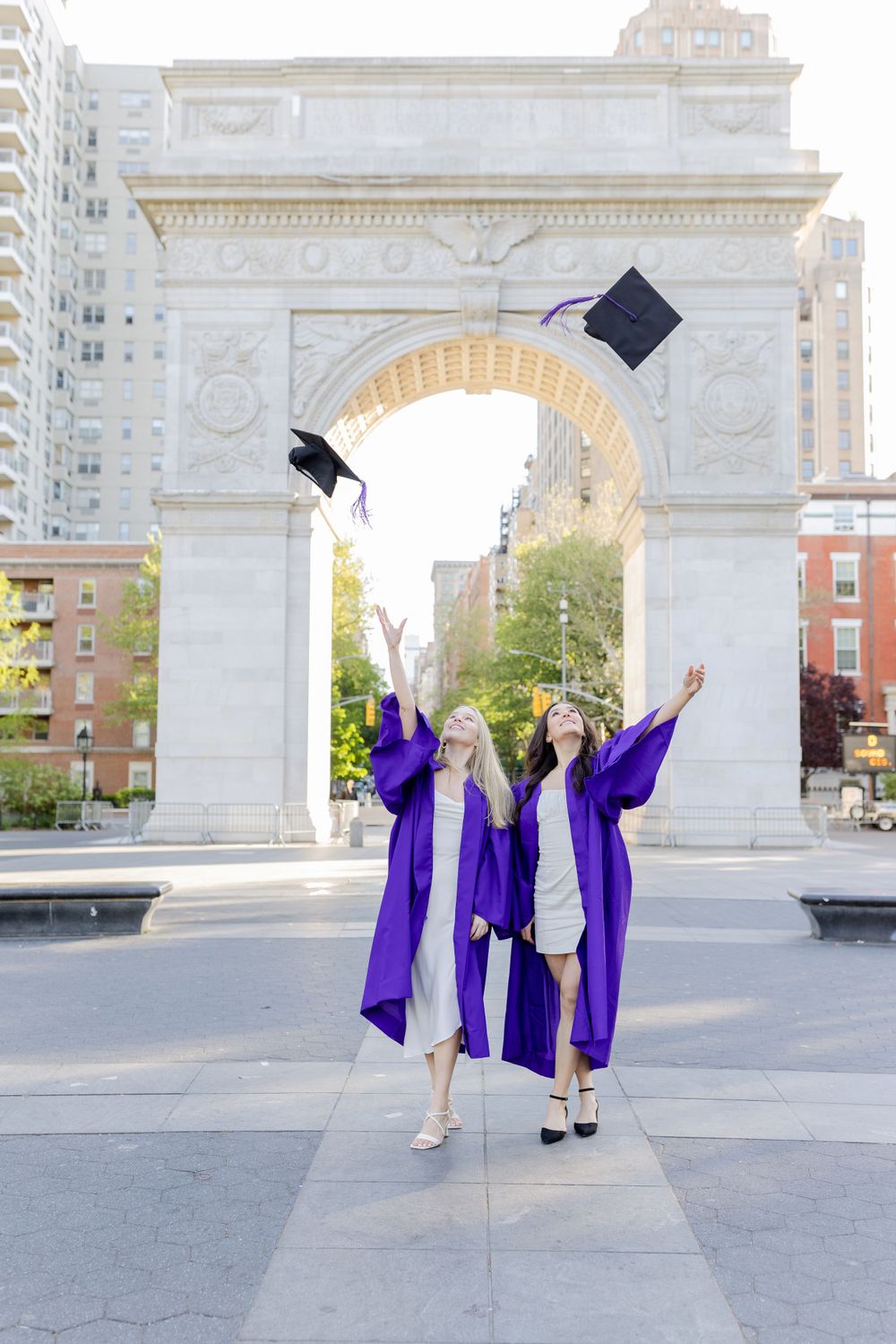 Graduates toss caps in celebration under Washington Square Arch wearing purple graduation gowns.