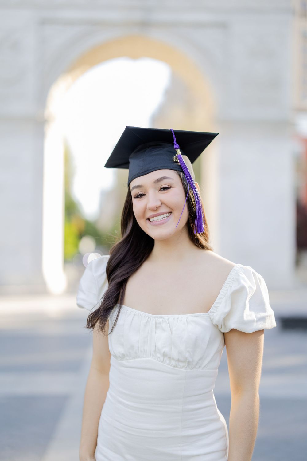 Graduate in white dress and cap poses in front of blurred architectural background.