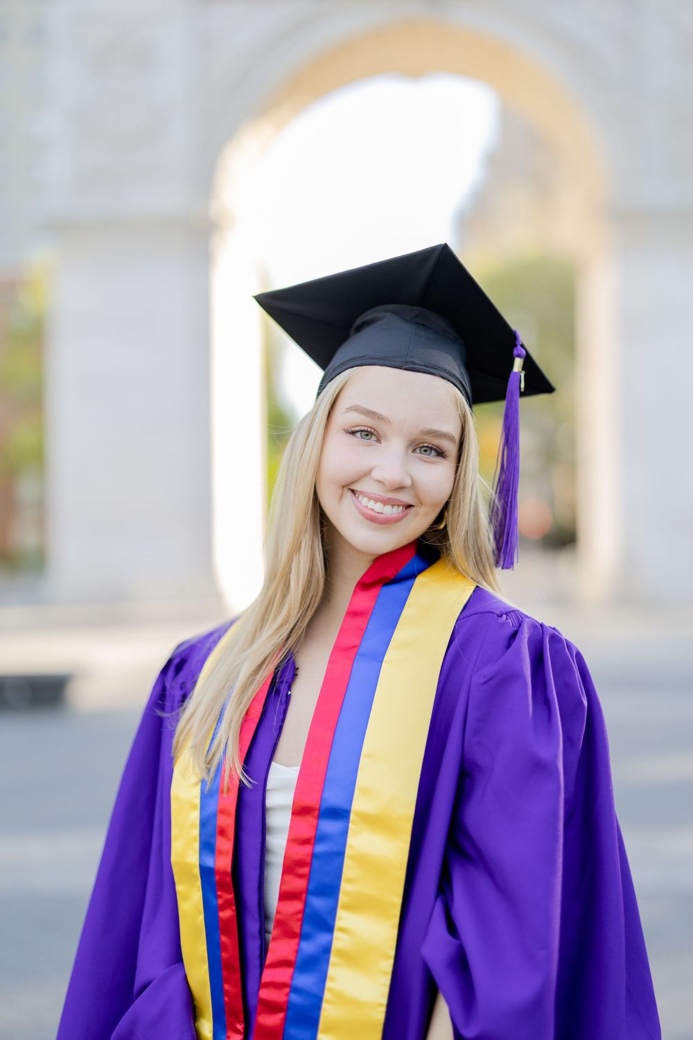 Graduate wearing purple gown with honors cords poses by historic arch.