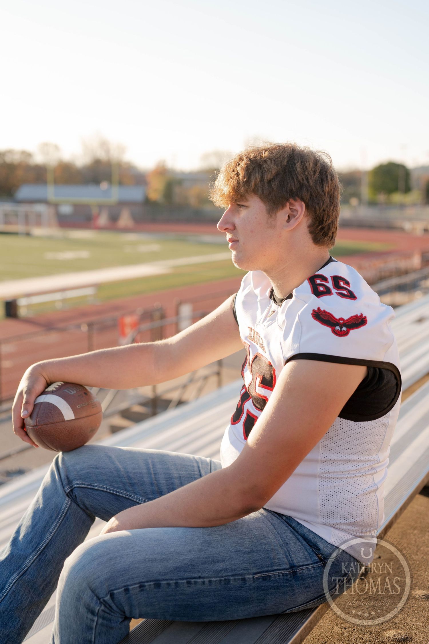 Football player in white jersey number 65 sits casually on stadium bleachers holding a football.