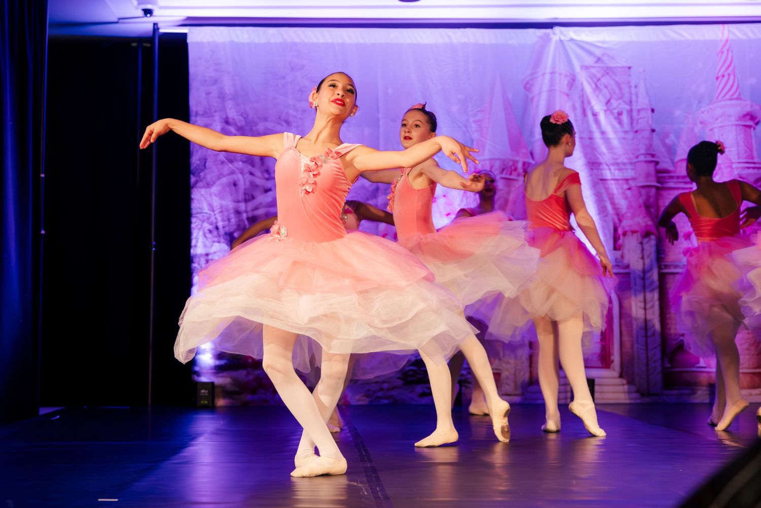 Ballet dancers in white tutus perform on stage under dramatic purple lighting during a classical dance recital.