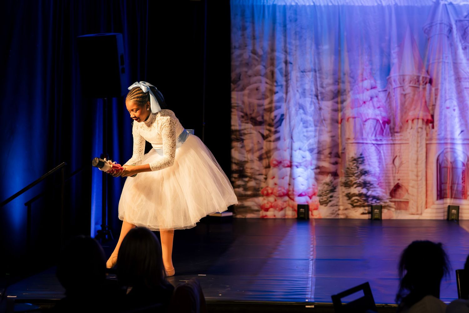 A ballet dancer in a white tutu dress performs on stage with dramatic blue lighting and scenic backdrop.