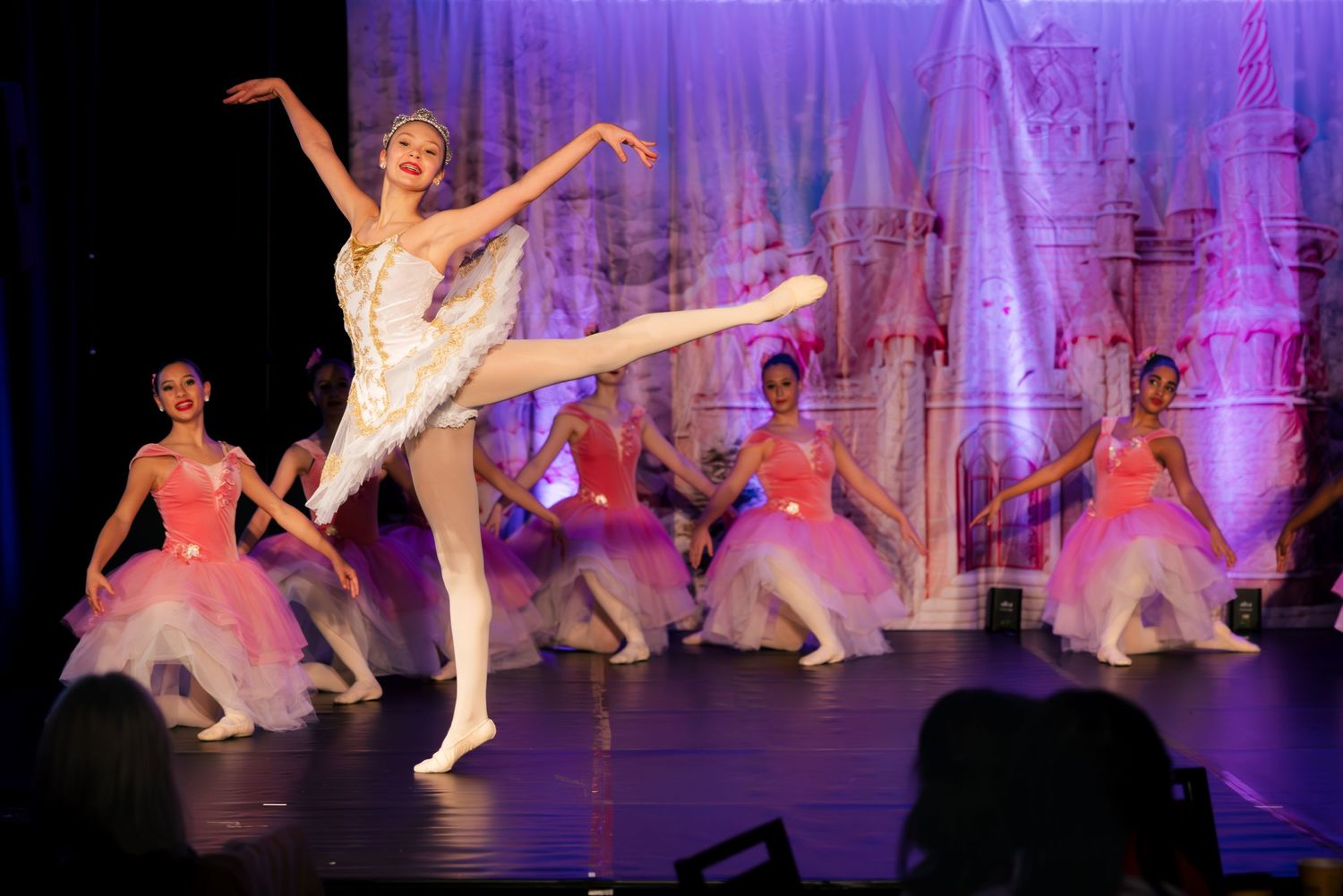 Ballet dancers perform on stage in white and pink tutus during a classical dance recital with purple lighting effects.