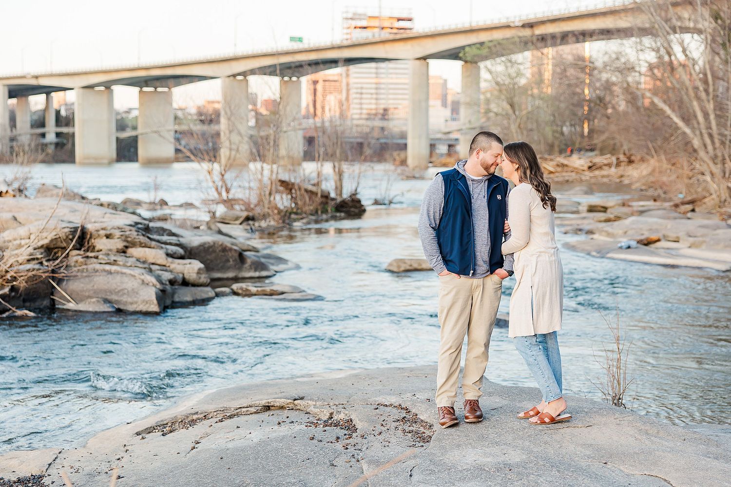 A couple stands close together on rocky terrain near a river with a bridge visible in the background during sunset.