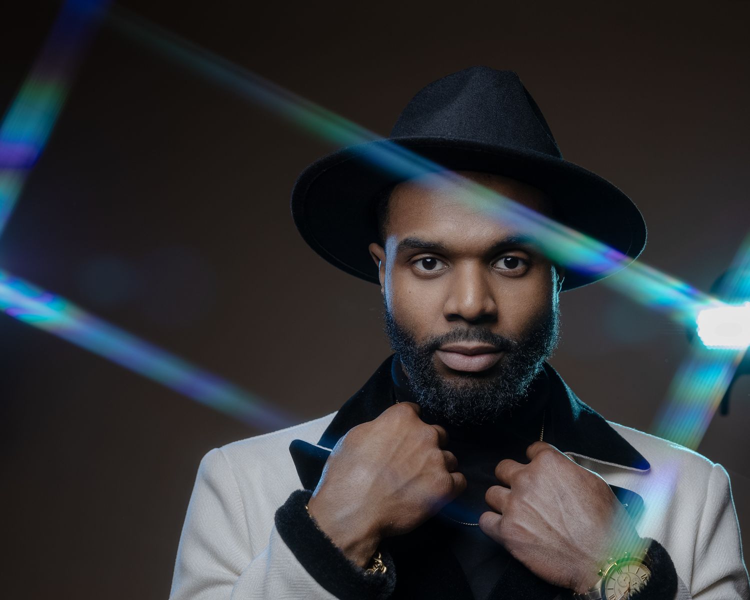 Stylish man in black hat and white jacket poses with hands on beard against dark background with blue lighting effects.