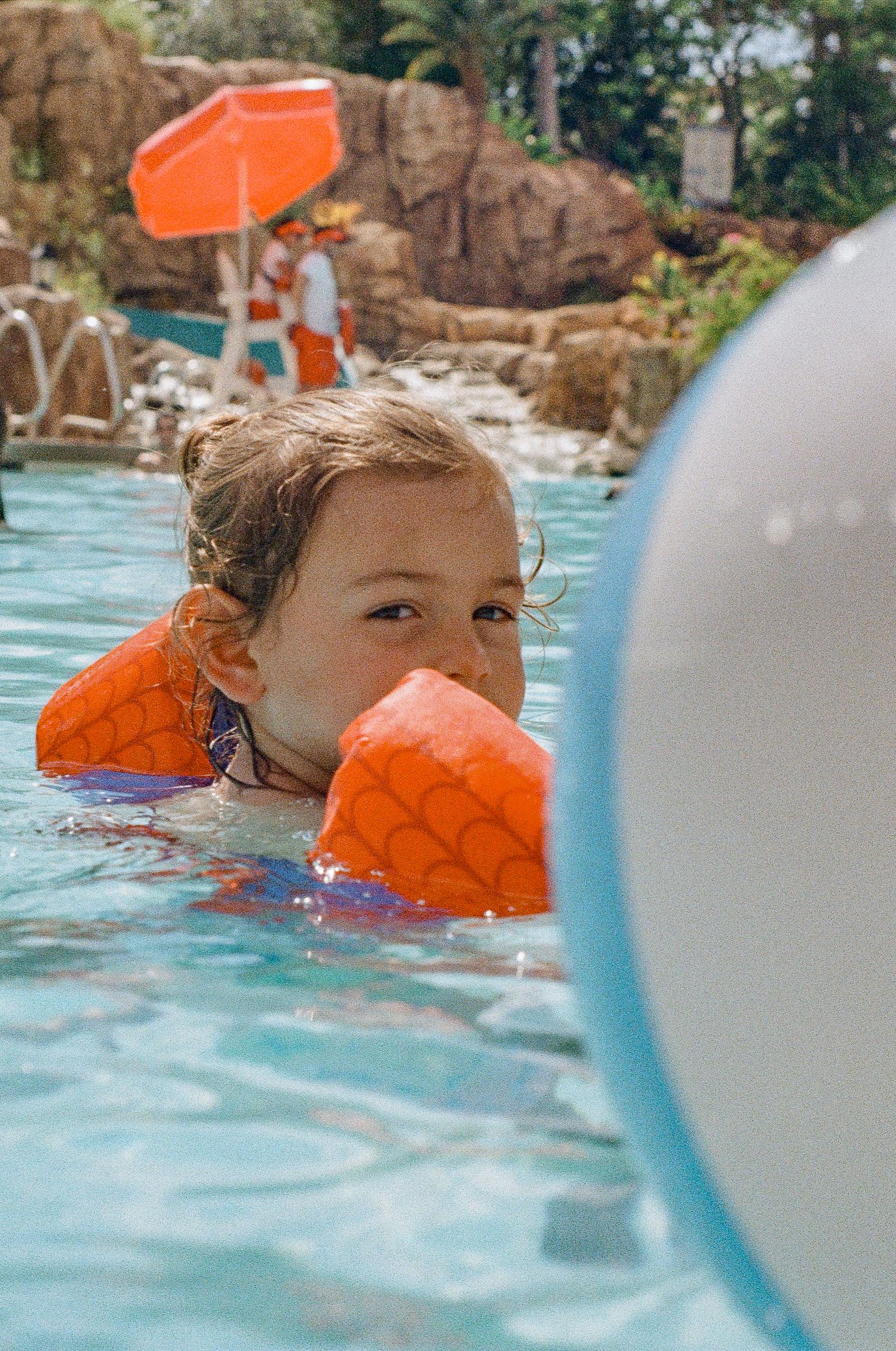 Someone swimming in a pool wearing orange arm floaties with a blue pool toy visible in the foreground.