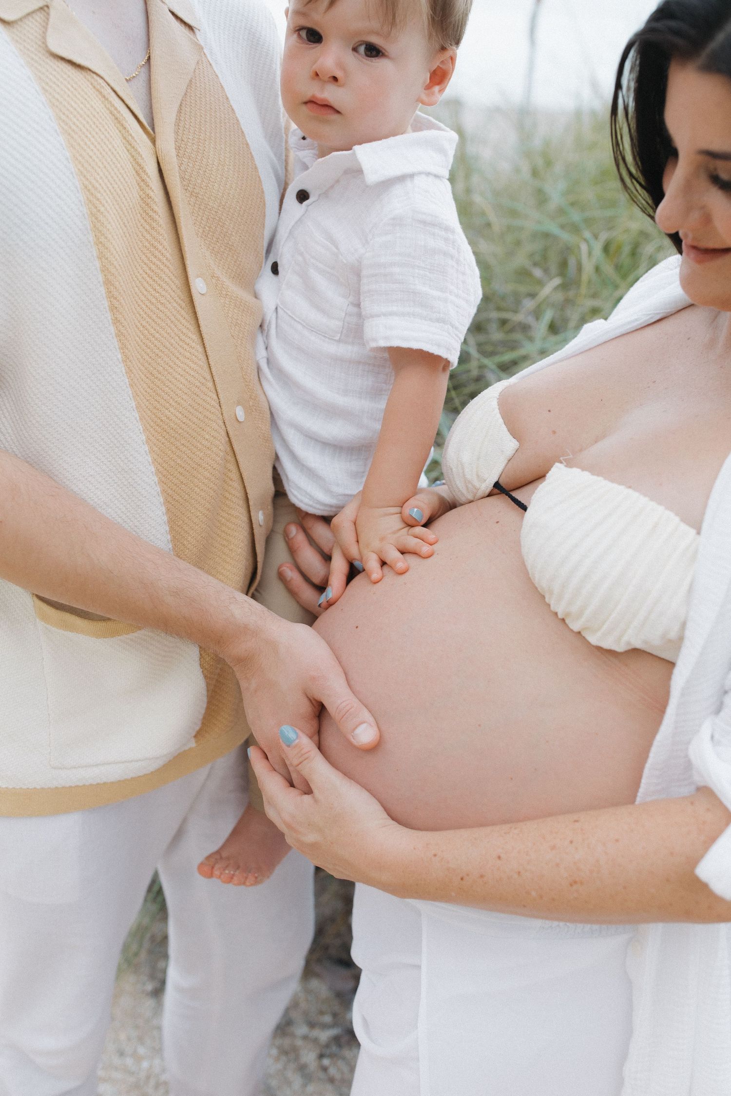 Pregnant belly shown in casual maternity portrait with light blue button-down shirt among tall grass outdoors.