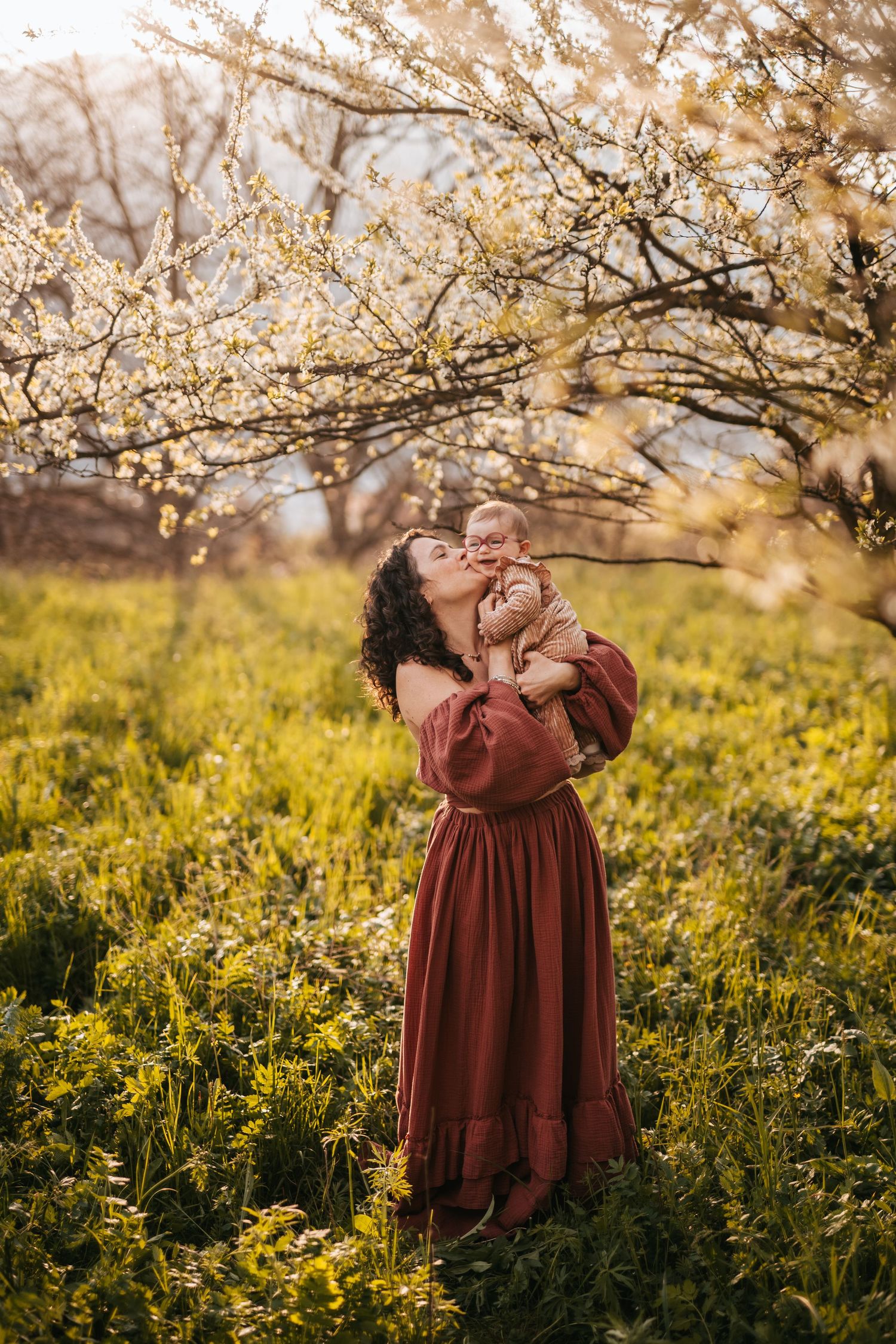 Giovane mamma tiene in braccio e bacia la sua bambina in un prato verde vicino a un albero in fiore a primavera
