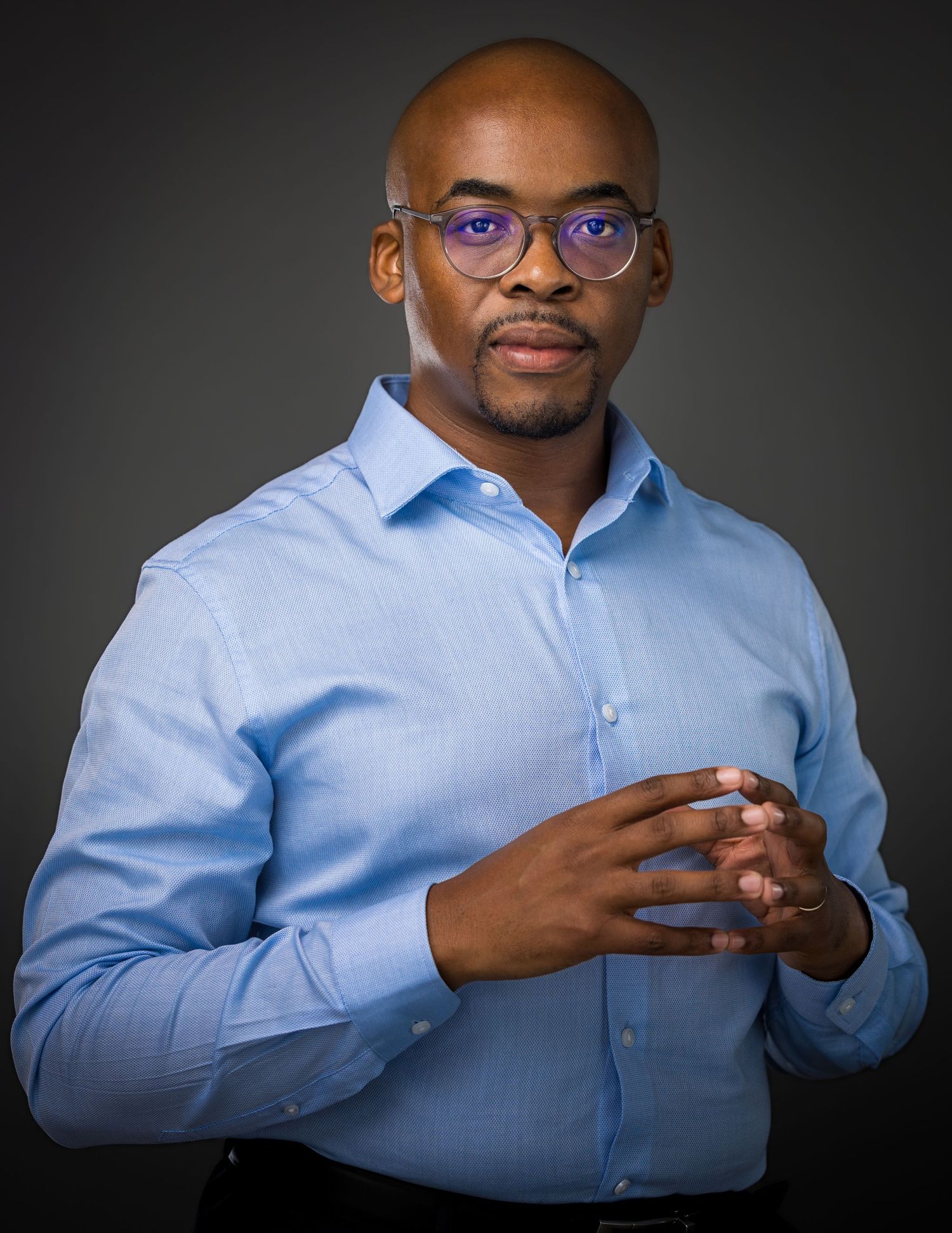 Professional headshot of someone in business attire with glasses, wearing white shirt and dark vest against black background.