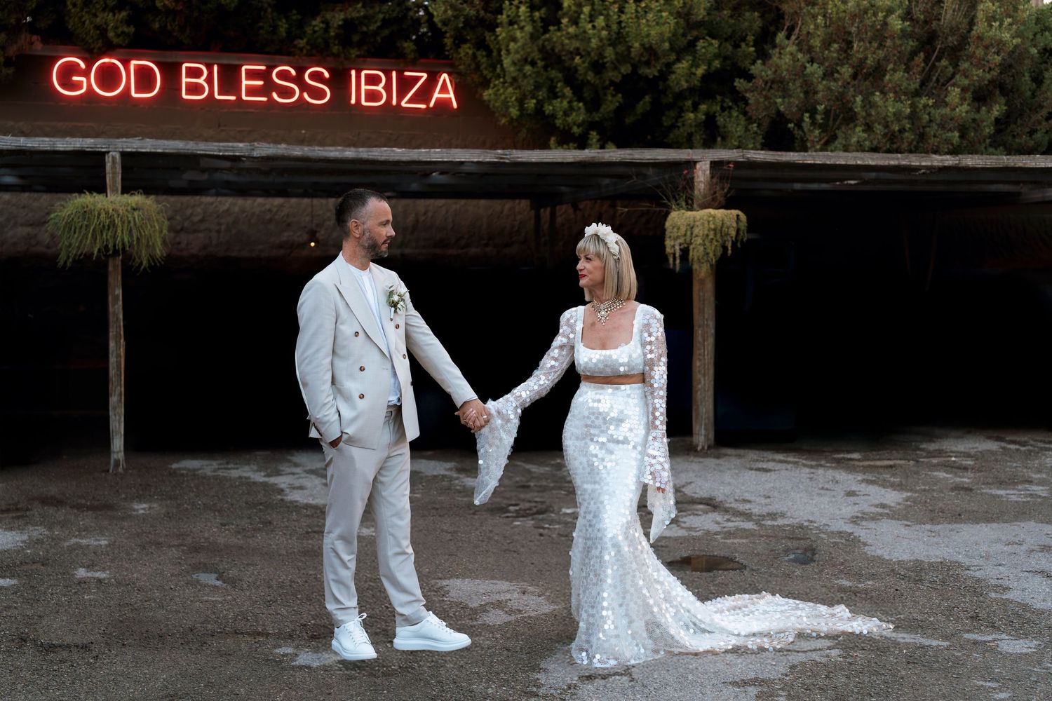 A couple in white wedding attire holds hands beneath a red neon 'God Bless Ibiza' sign.