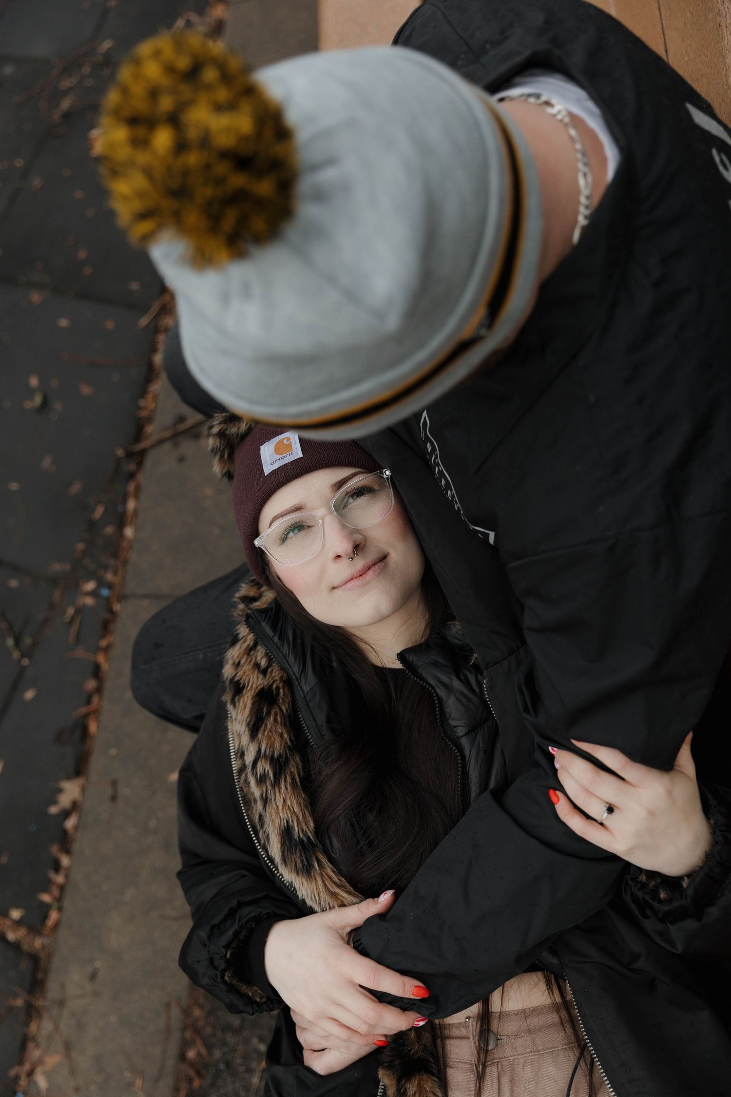Person wearing a black coat and beanie with a yellow pom pom lies on concrete near fallen leaves in moody lighting.