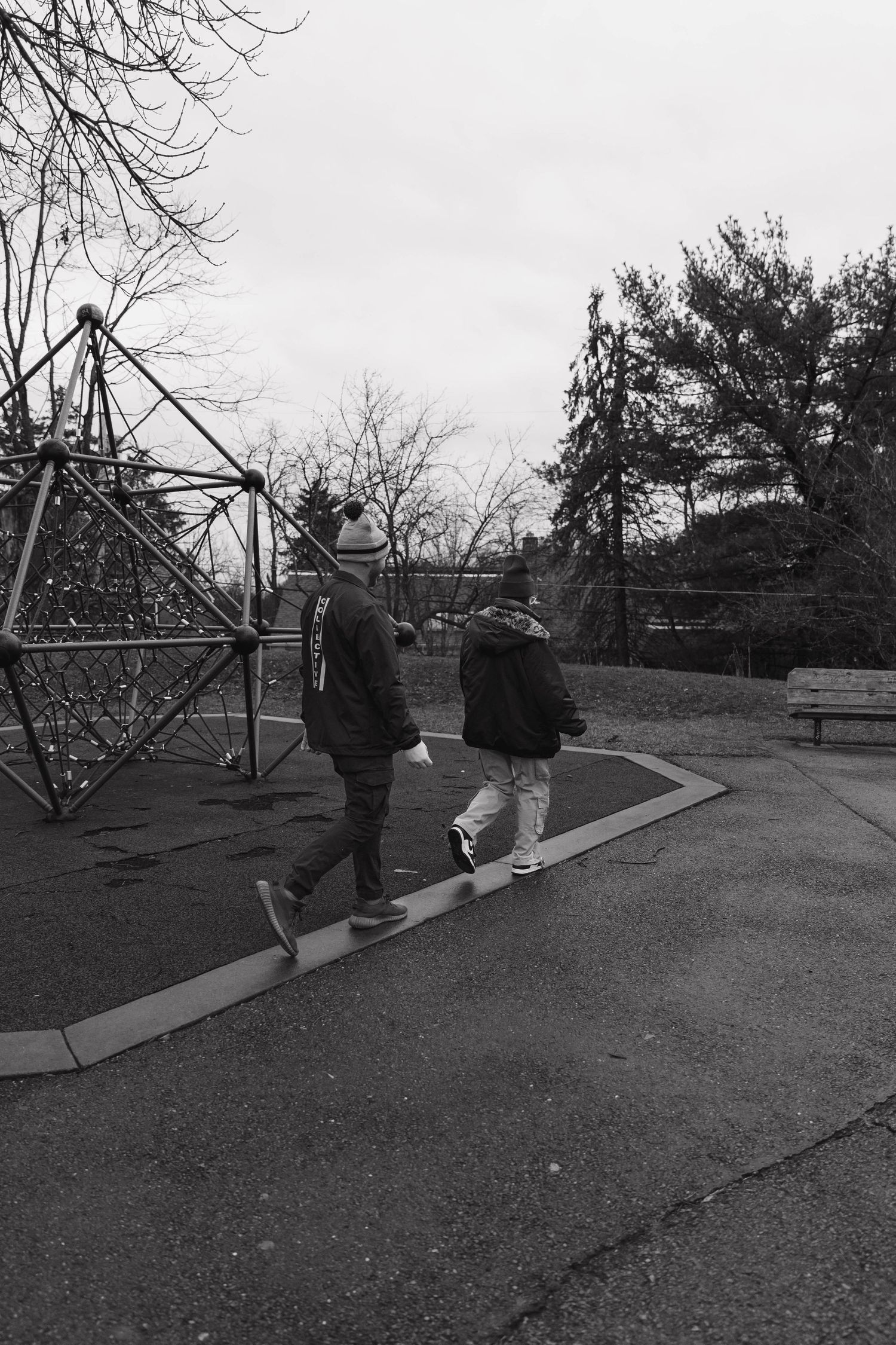 Two people walking in a park with playground equipment and bare winter trees in black and white photography.