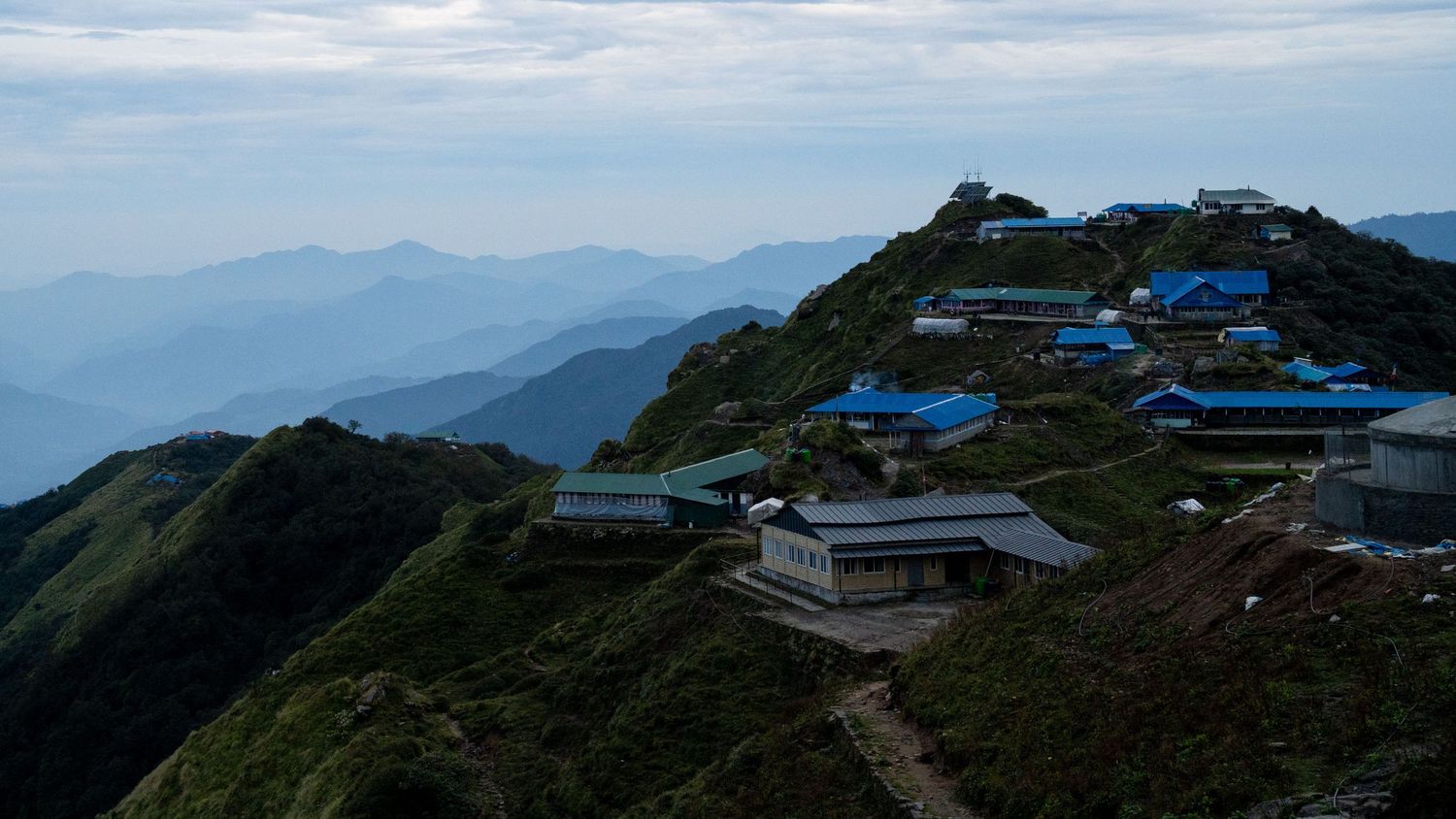 Mountain village with blue-roofed buildings perched on terraced hillside overlooking valley.