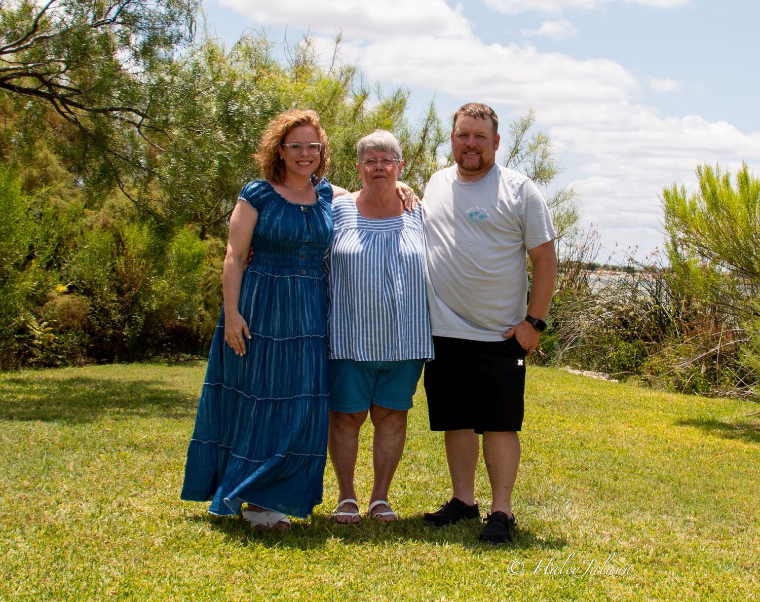Three people posing together outdoors on a sunny day with green trees and grass in the background.