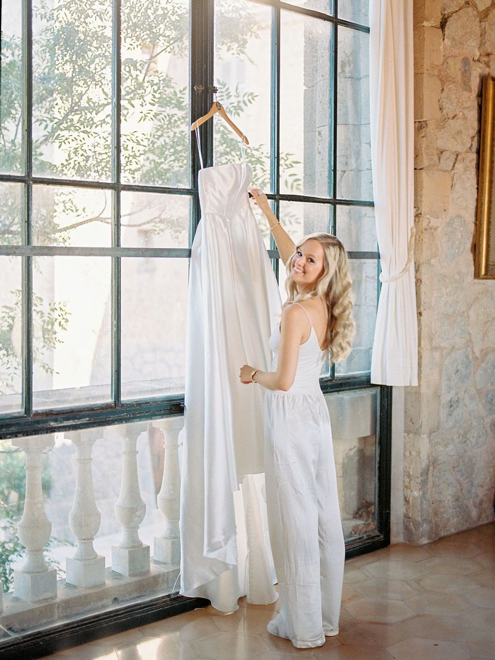 Wedding dress hanging by large windows in an elegant getting ready room.