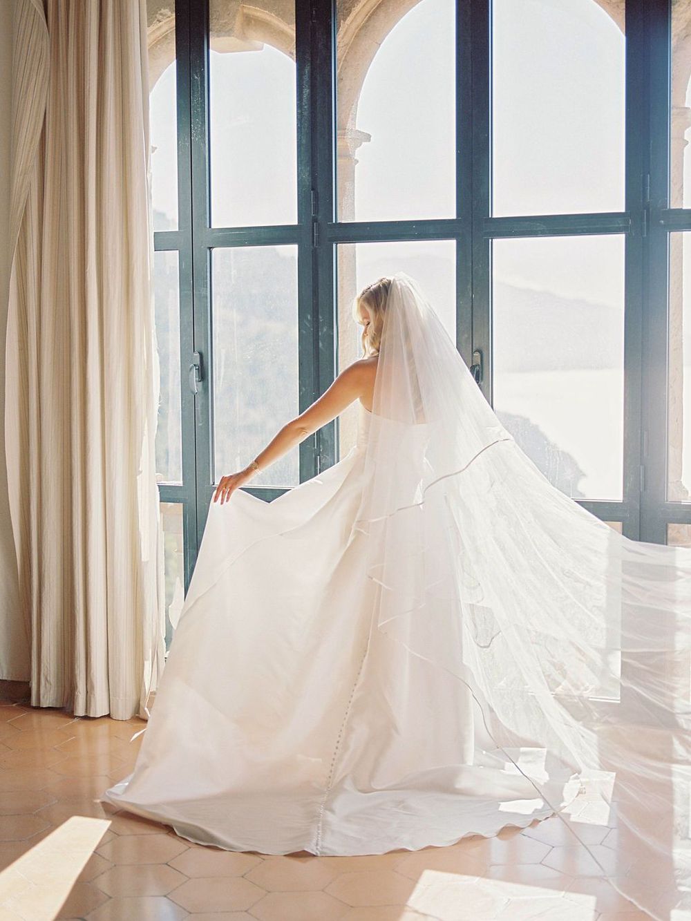 Flowing wedding dress with long train and veil photographed against bright windows.