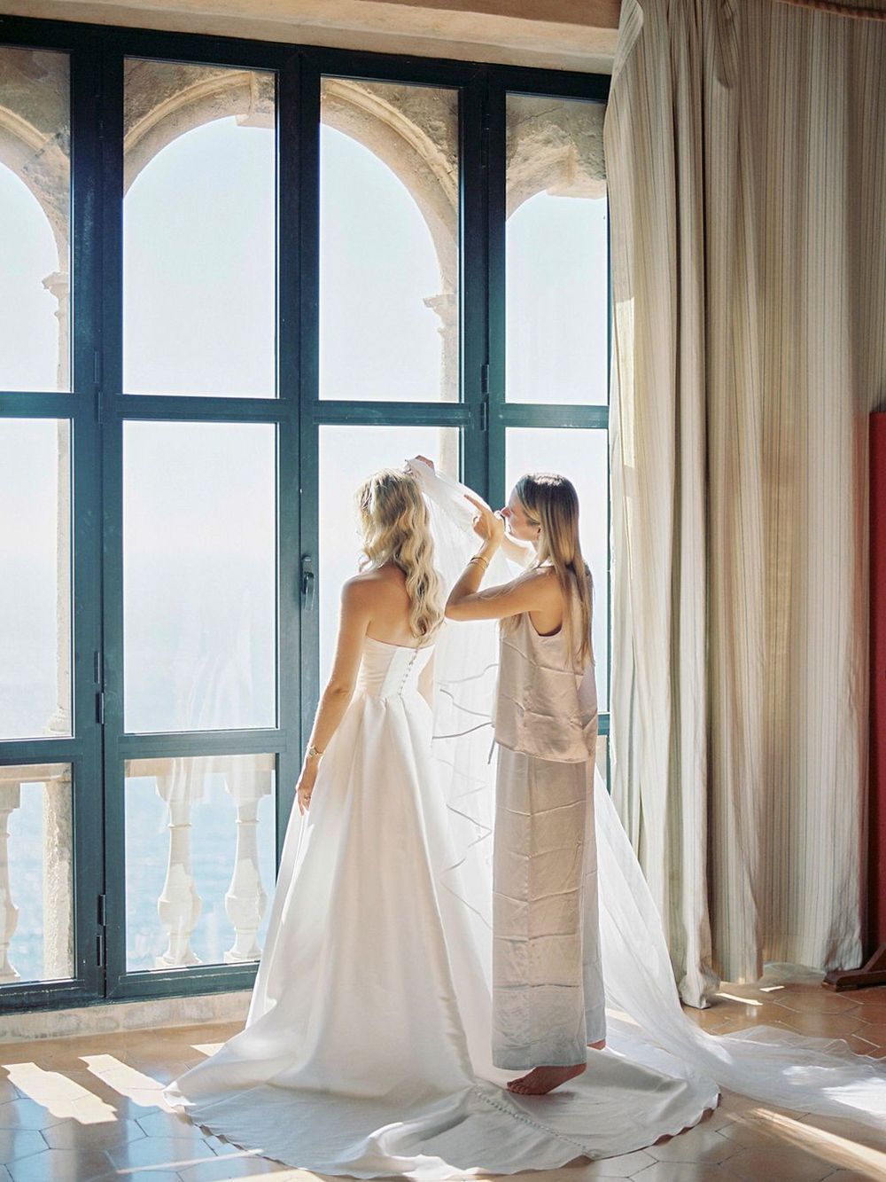 Wedding preparations by a sunlit arched window in an elegant stone interior with blue shutters.