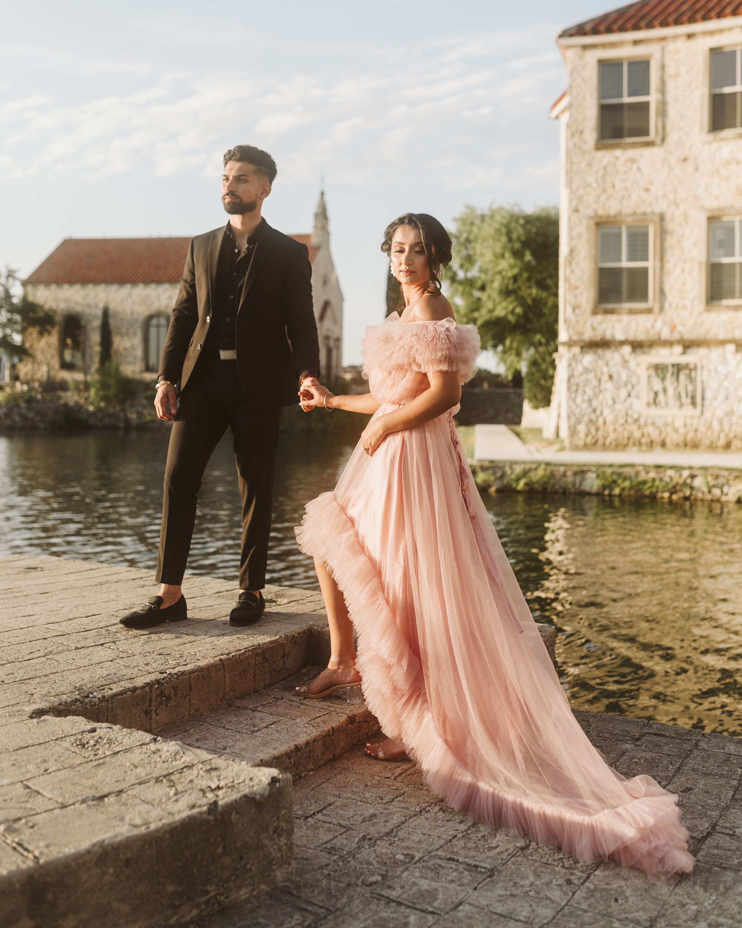 Couple in formal wear poses along a waterfront with pink flowing gown and European architecture.