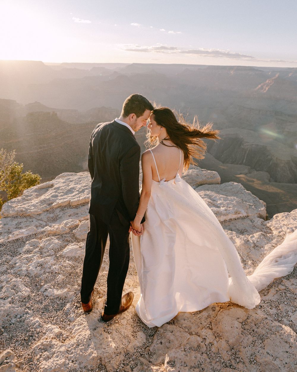 Couple stands at edge of Grand Canyon during sunset in wedding attire with dress flowing in wind.