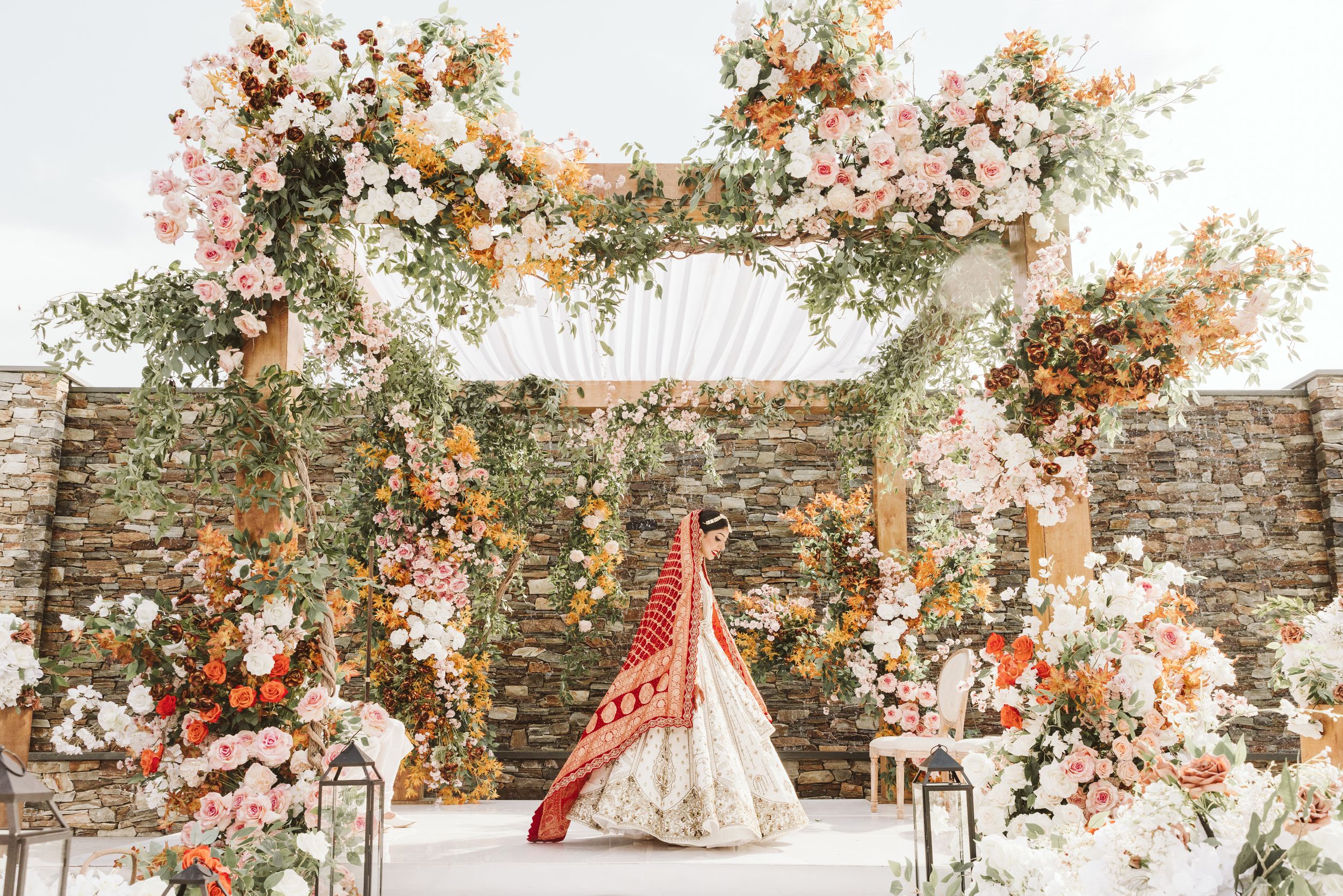 Elegant wedding arch adorned with coral and cream flowers against rustic stone wall with romantic flowing dress.