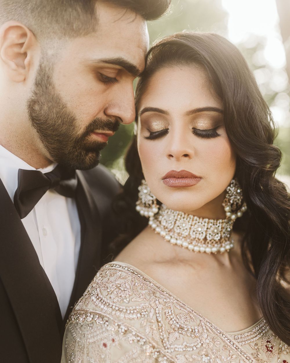 Elegant South Asian couple in wedding attire with ornate jewelry and makeup in soft lighting.