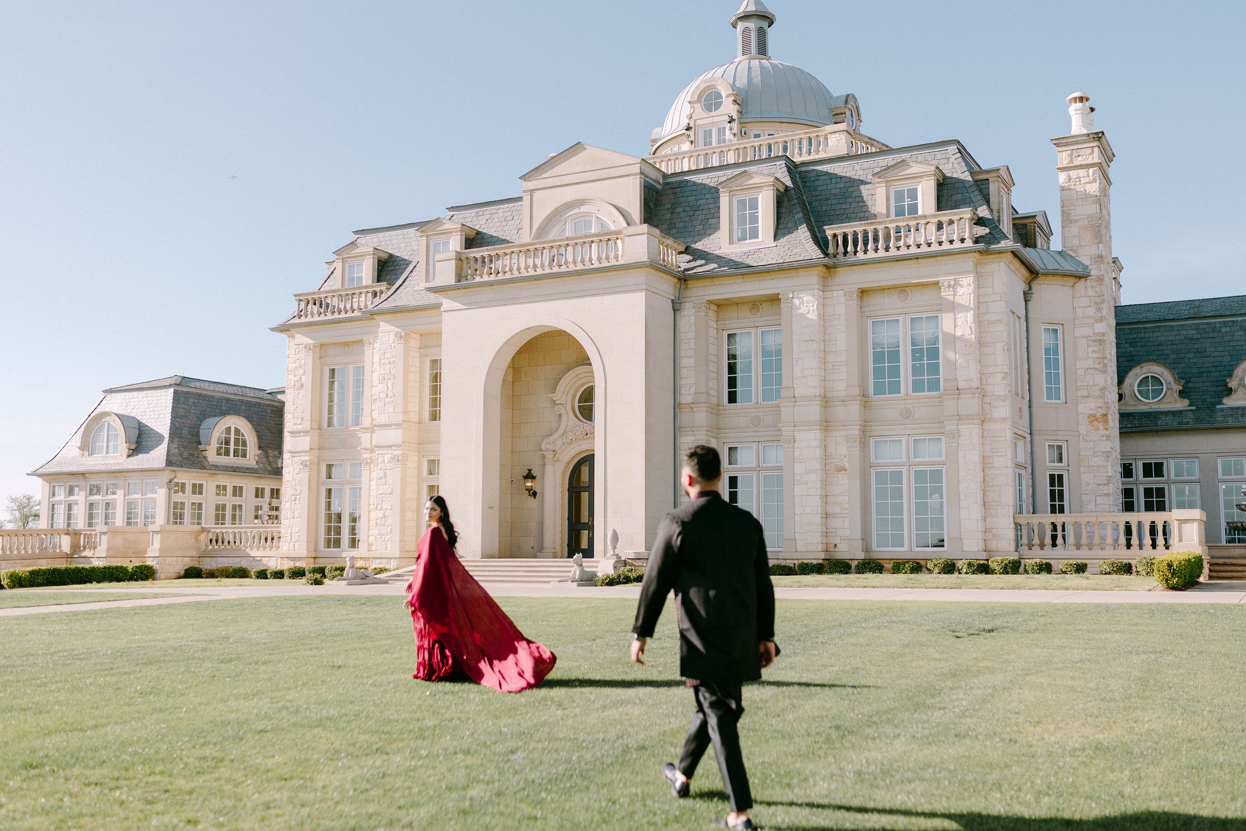 Two people walk outside an elegant domed mansion with white stone facade and grand archway on a sunny day.