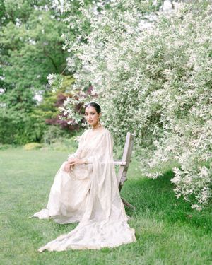Ethereal bridal portrait in a flowing white dress against blooming spring trees in a garden setting.