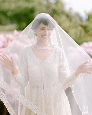 A bride wearing a sheer veil and white lace dress smiles joyfully while standing in front of pink flowers outdoors.