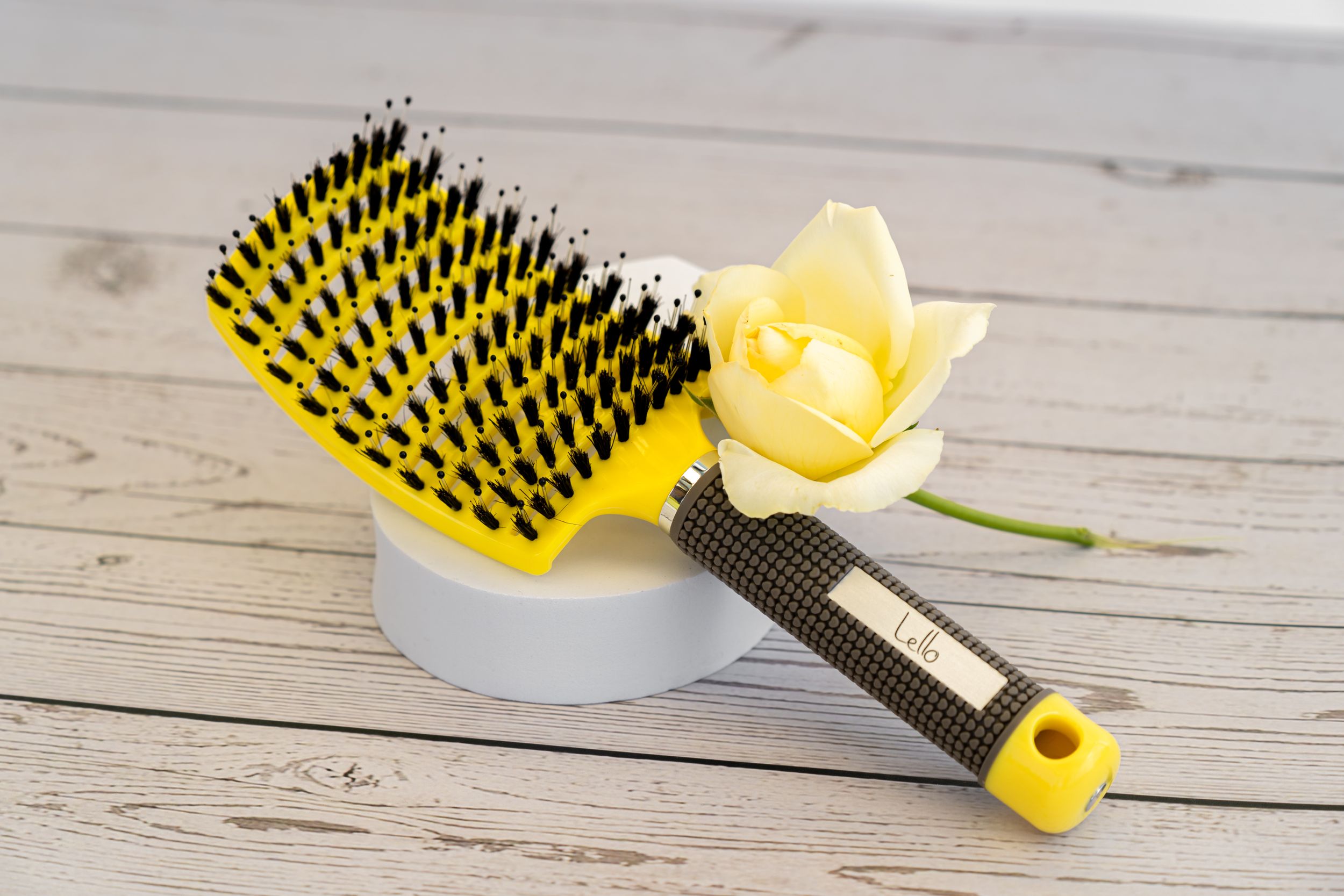 Styling brush with yellow bristles next to fresh daffodil on white wooden surface.