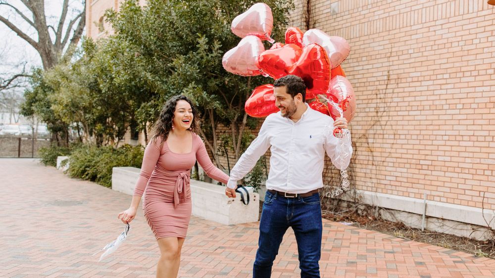 San Antonio Maternity Photography. A couple in pink and white attire walks down a brick path holding red heart balloons.