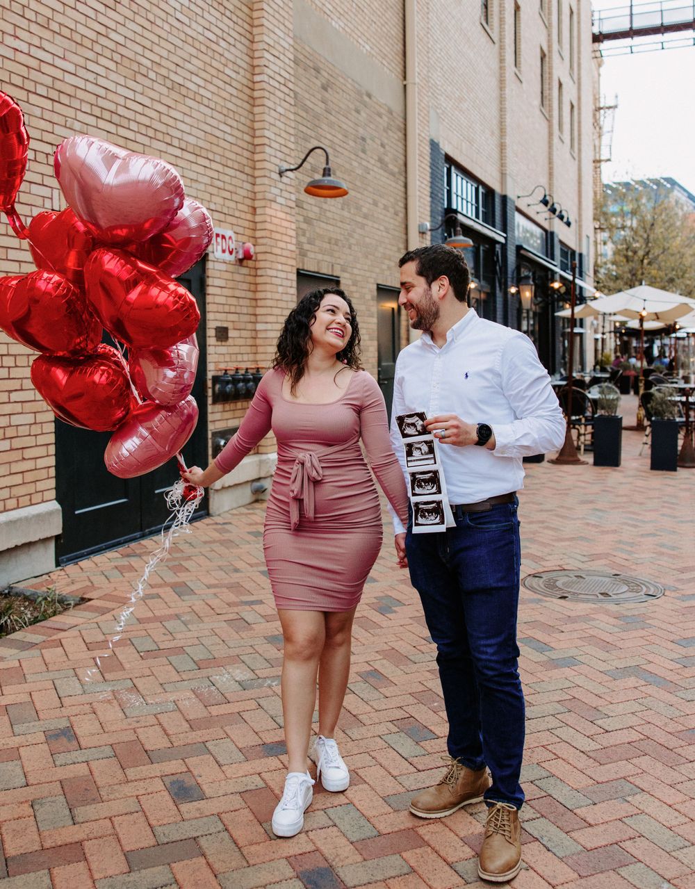 San Antonio Maternity Photography. A couple walks down a city street holding heart balloons and ultrasound photos.