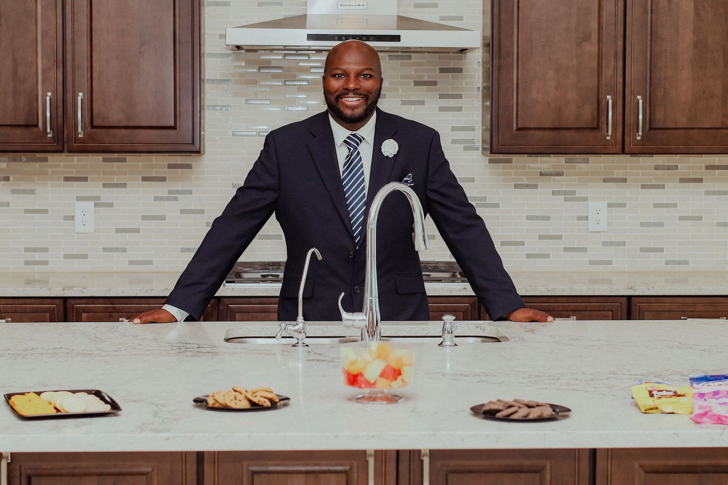 A smiling man in a navy suit stands behind a kitchen island with snacks and treats.