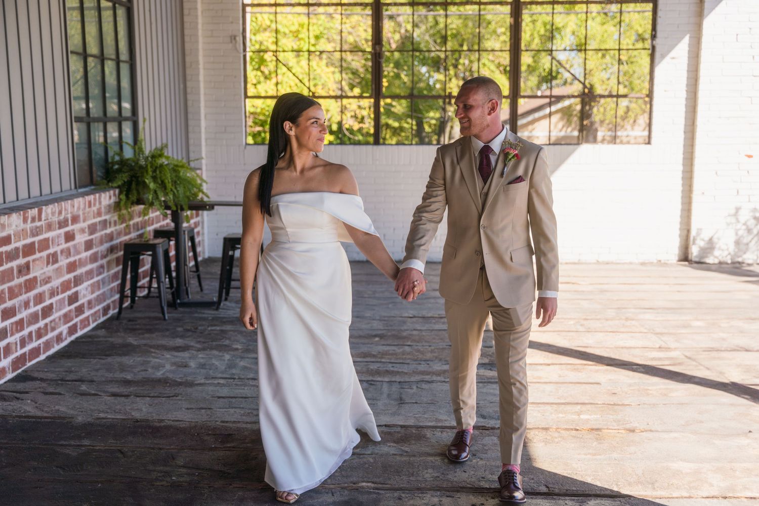 A couple in wedding attire walks hand in hand through a bright indoor venue with large windows.